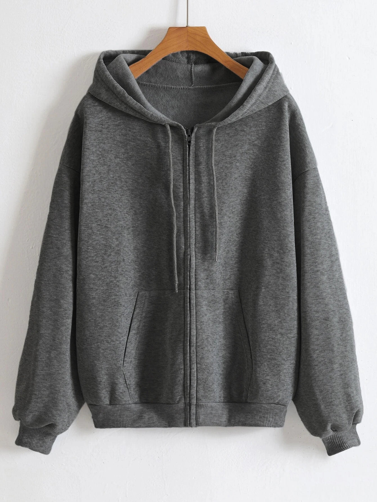 A gray oversized zip-up hoodie hanging on a wooden hanger against a white wall.