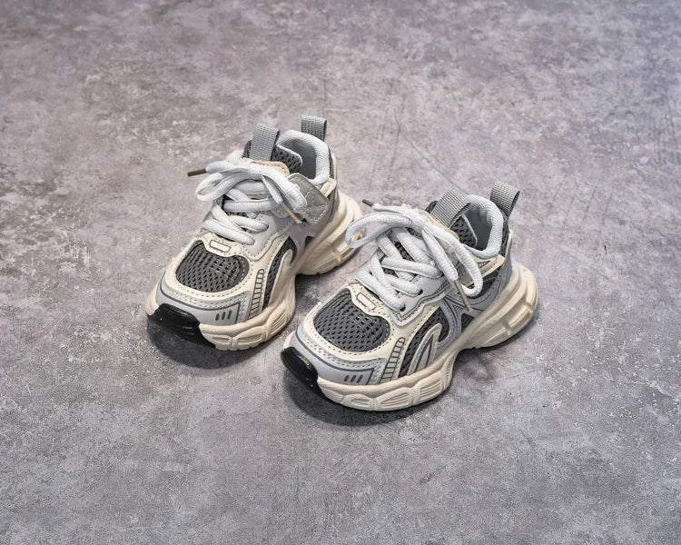A pair of gray and white breathable mesh kids running sneakers with cushioned soles resting on a gray concrete surface.