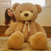 A large, light brown teddy bear plush toy sitting on a beige carpet next to a young woman.