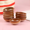 A set of ten miniature brown rice bowls arranged in a stack on a pink surface.