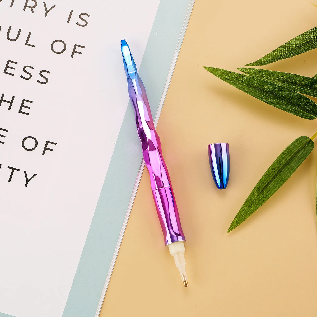 A purple diamond painting pen is shown next to a green leaf on a light beige background.