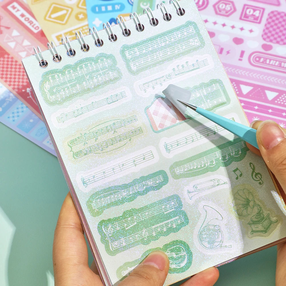 A hand holds an open sticker book filled with sheet music and decorative elements for fantasy aesthetic journaling.