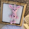 A handmade crochet frog hanging ornament in pink with black eyes displayed in a wooden frame.