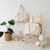 Three cream quilted diaper bags with floral embroidery displayed on a wooden chair against a white wall.