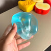 A hand holds a translucent blue squishy stress relief fidget toy next to yellow and red fruit shapes.