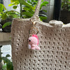 A handmade crochet pink dinosaur keychain hanging from a natural fiber tote bag.