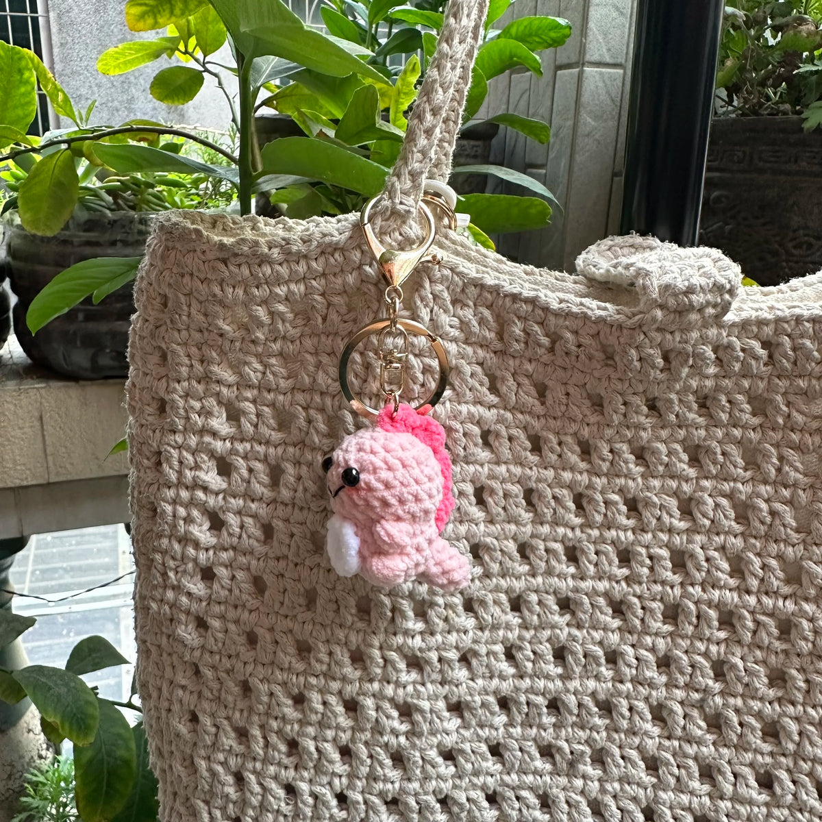 A handmade crochet pink dinosaur keychain hanging from a natural fiber tote bag.