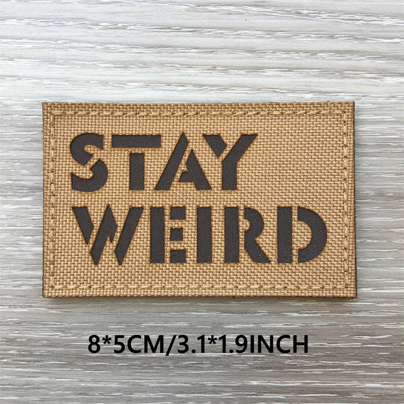 Un patch en tissu beige rectangle avec du texte noir disant STAY WEIRD, posé sur une surface en bois clair.
