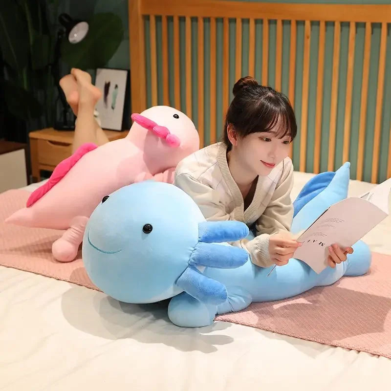 A young woman is sitting on a bed surrounded by large colorful axolotl plush pillows.