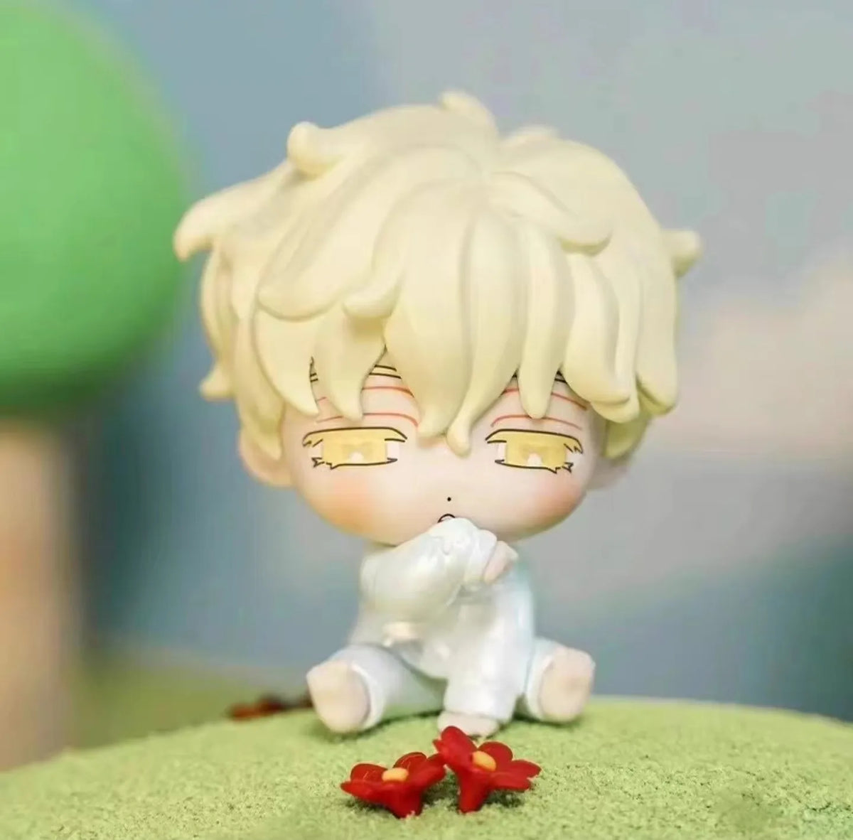 A small, pale blonde mini figure collectible with large eyes and white clothing sits on a green surface.