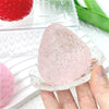 A hand holds a pink, translucent strawberry squishy toy in a clear dish next to a red fruit.