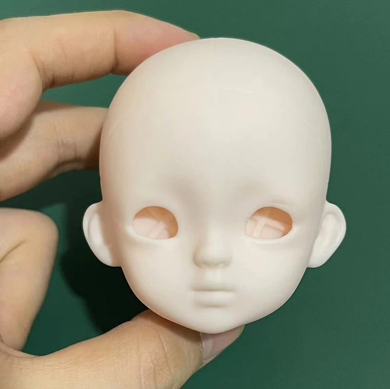 A hand holds a smooth, pale pink 1/6 scale BJD doll with large eyes against a dark green background.