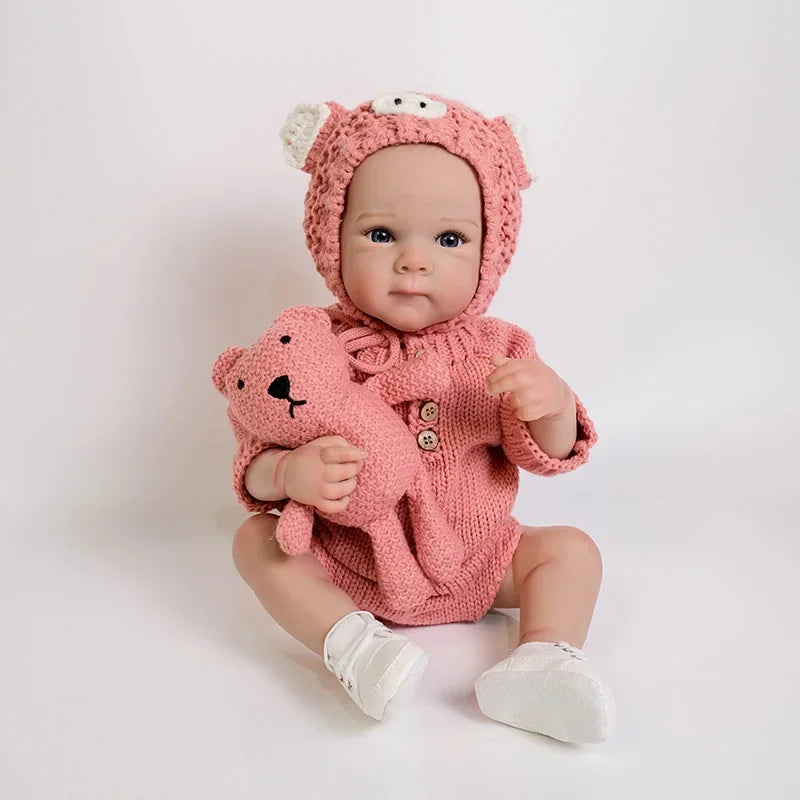 A realistic silicone reborn baby doll wearing a pink knitted outfit and hood, sitting on a white surface.