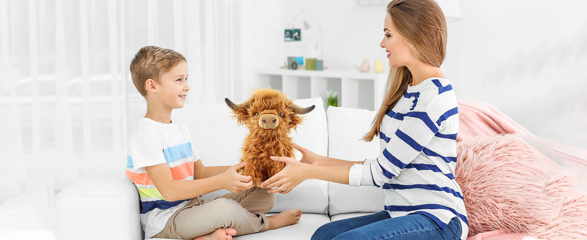 Eine Frau zeigt einem Jungen einen flauschigen braunen Highland-Kuh-Plüschtier auf einem weißen Sofa.