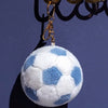 A soft stuffed soccer ball toy with white and blue patches is hanging from a gold-colored chain.