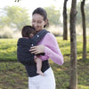 A woman wearing a pink top is holding a baby in an adjustable cotton carrier wrap outdoors.