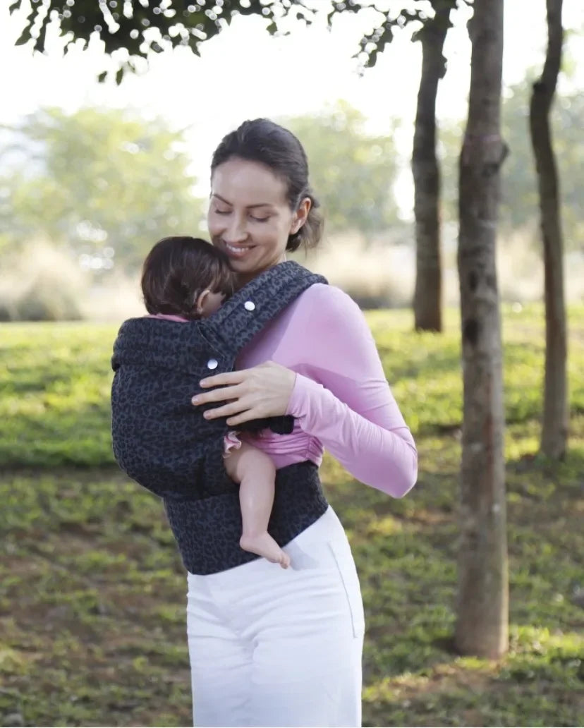 A woman wearing a pink top is holding a baby in an adjustable cotton carrier wrap outdoors.