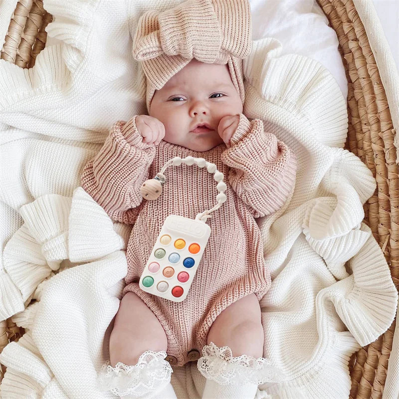 A baby wearing a knitted outfit and hat is holding a silicone teether toy shaped like a cartoon car.