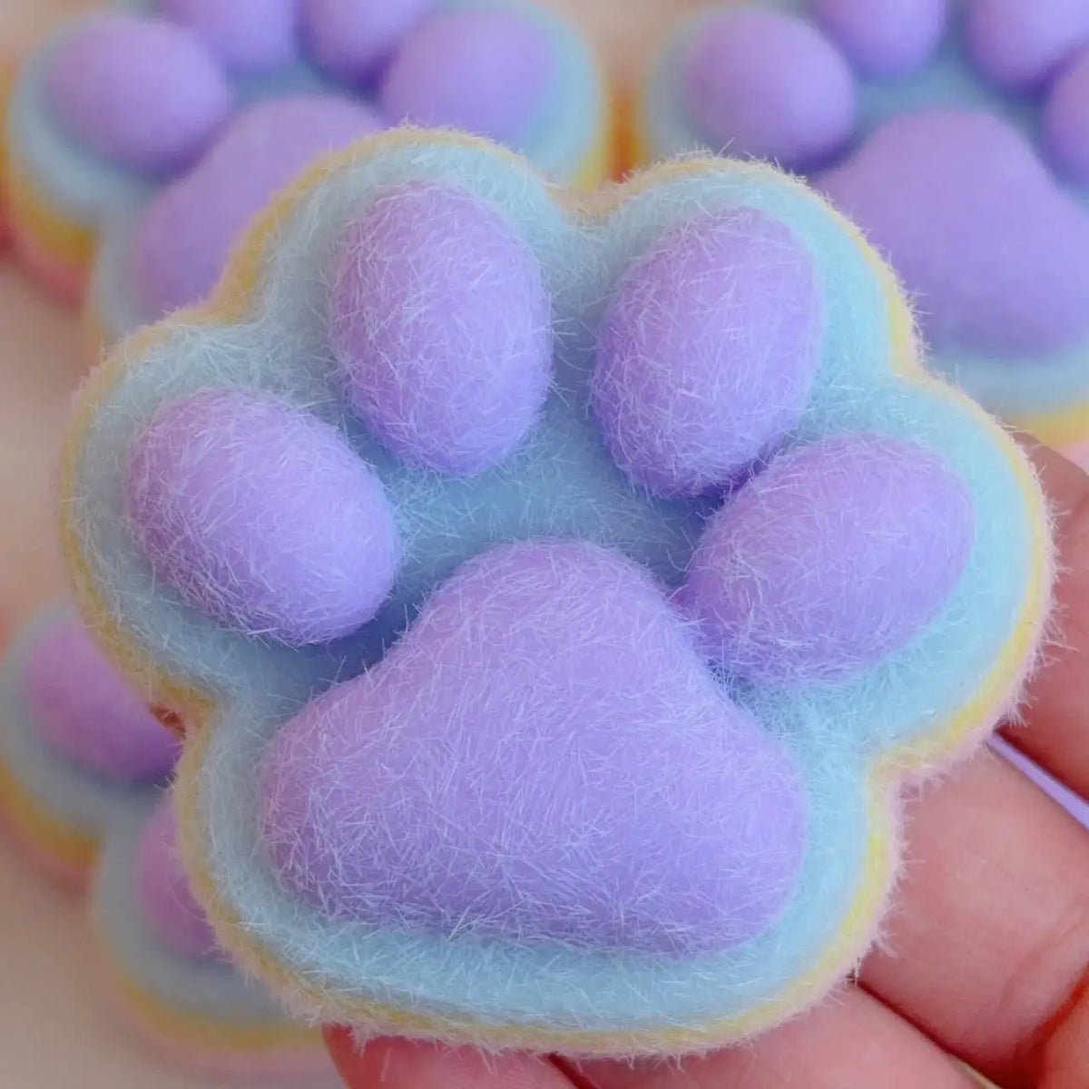 A hand holds a soft, purple cat paw shaped squishy fidget toy with a light blue rim.