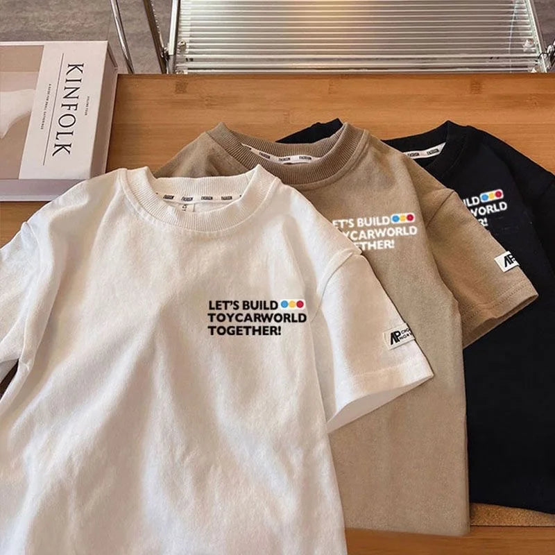 A close-up of a white kids car print crew neck t-shirt lying next to two other colored shirts.