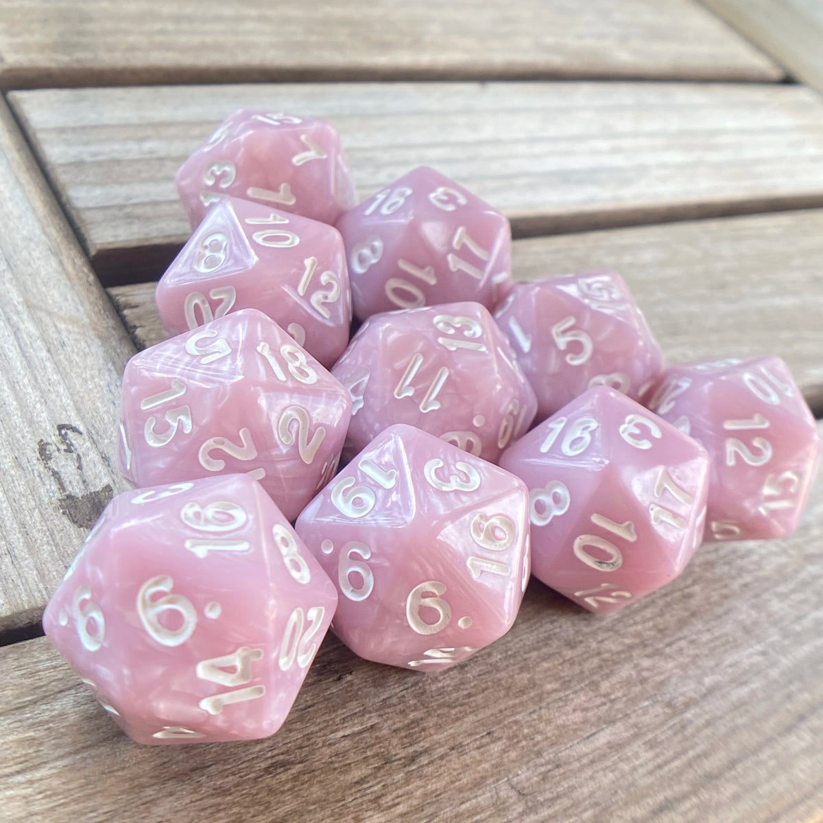 A set of ten pink pearl acrylic D20 dice arranged in a row on a wooden surface.