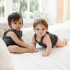 Two toddlers wearing dark denim rompers sitting on a white couch near a window.