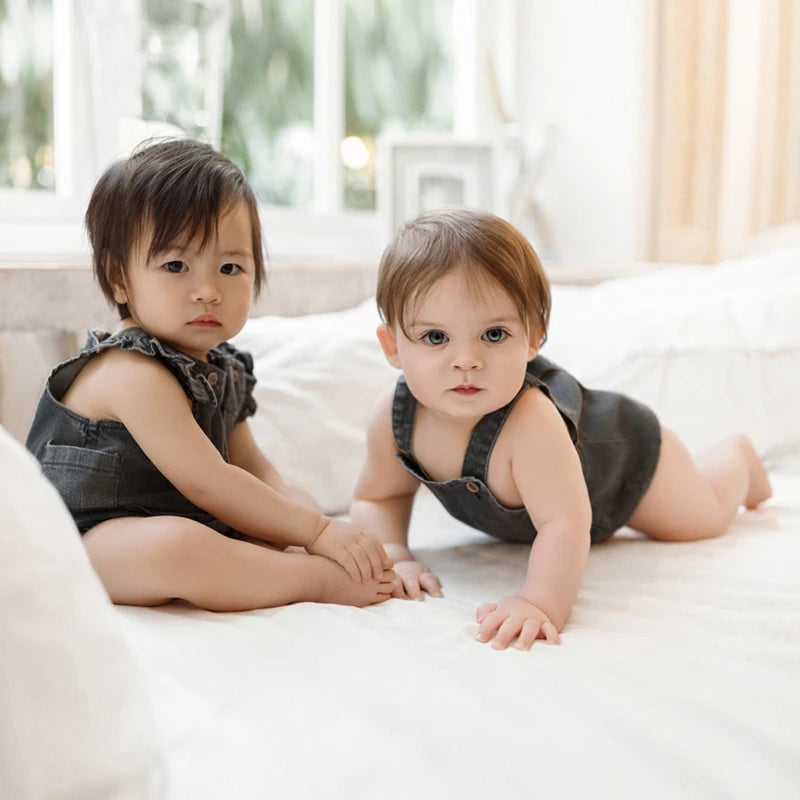 Two toddlers wearing dark denim rompers sitting on a white couch near a window.