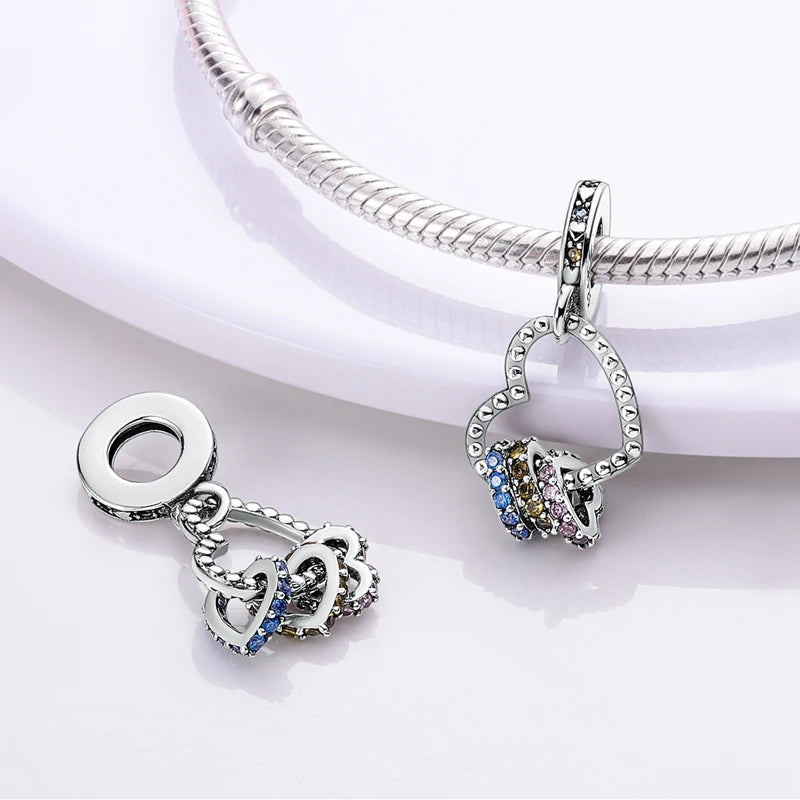 A silver heart charm with multicolored stones is displayed on a silver chain and next to a separate pendant.