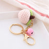 A pink crocheted fruit pendant keychain with a green stem attached to a gold ring on a white plate.