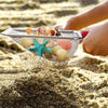 A hand is scooping seashells and starfish from the sand using a metal mesh scoop.