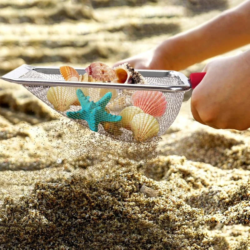 A hand is scooping seashells and starfish from the sand using a metal mesh scoop.