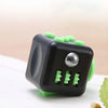 A black fidget cube with green buttons and a silver magnetic button, resting on a white surface.
