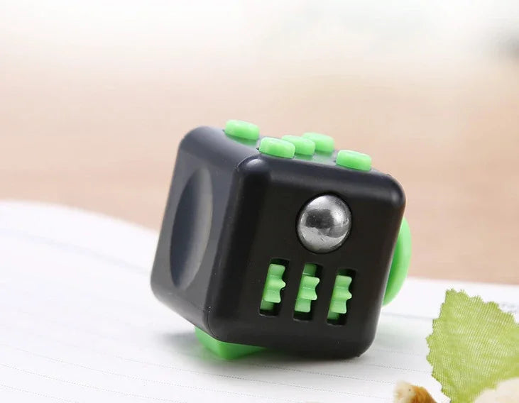 A black fidget cube with green buttons and a silver magnetic button, resting on a white surface.