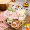 A plush keychain featuring a pink character with green accents and a small food item, displayed on a wooden tray.