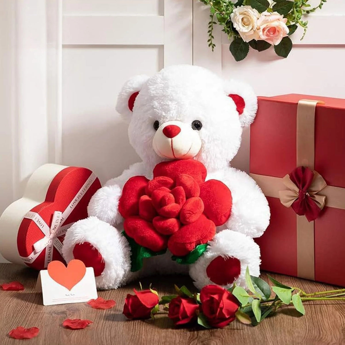 A white plush bear holding a red rose, set against a pink gift box and scattered rose petals.