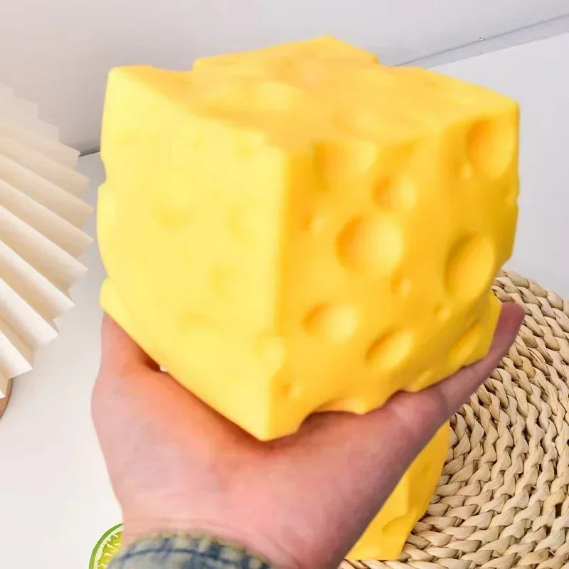 A hand holds a bright yellow cheese wedge squishy stress relief toy against a woven background.