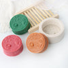 Three silicone molds shaped like faces in green, pink, and orange resting on a wooden tool and white fabric.