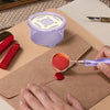 A hand is using a purple wax seal melting kit with a heater and spoon to melt wax on brown paper.