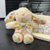 A light beige soft bunny plush toy with an orange and yellow floral bow charm resting on a dark surface.