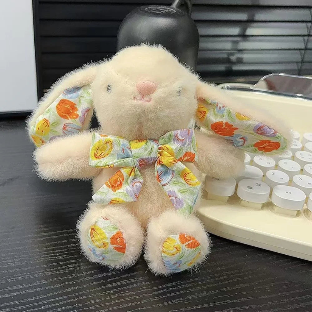 A light beige soft bunny plush toy with an orange and yellow floral bow charm resting on a dark surface.