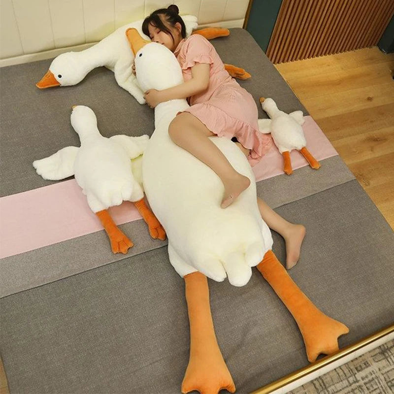 A woman is cuddling an extra-large white plush goose pillow with orange feet on a gray surface.
