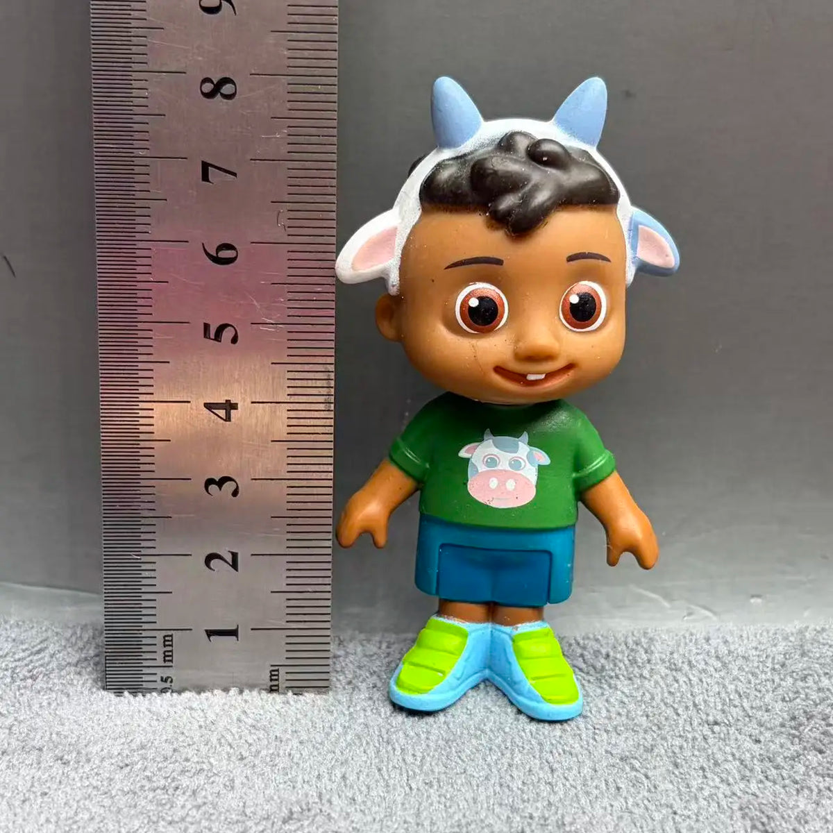 A playful cartoon PVC figurine of a child with dark skin, wearing a green shirt and blue shorts, against a ruler.