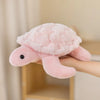 A soft pink sea turtle plush toy is being held by a person against a light background.
