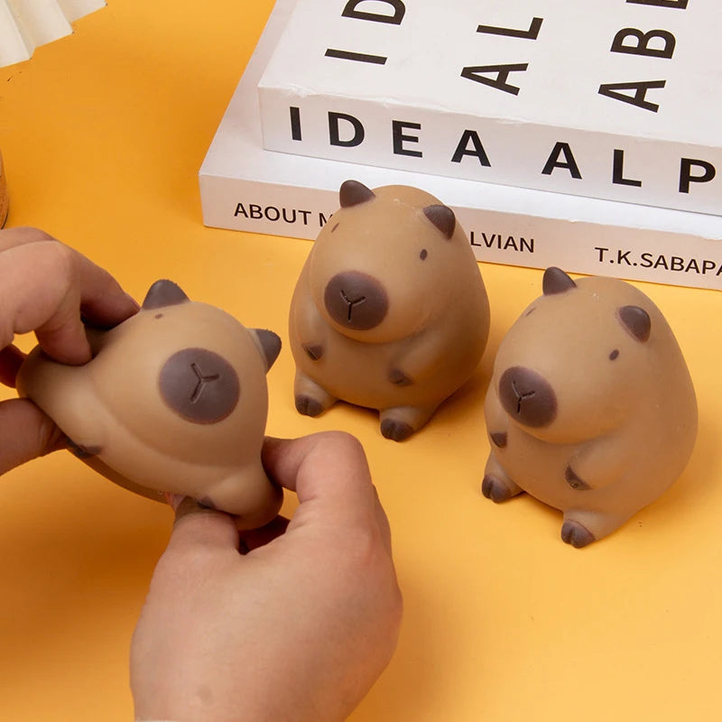 A hand holds a beige capybara squeeze stress relief toy next to a book.