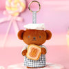 A brown teddy bear plush toy dressed as a baker with a checkered apron and a layered bread item, attached to a gold keyring.