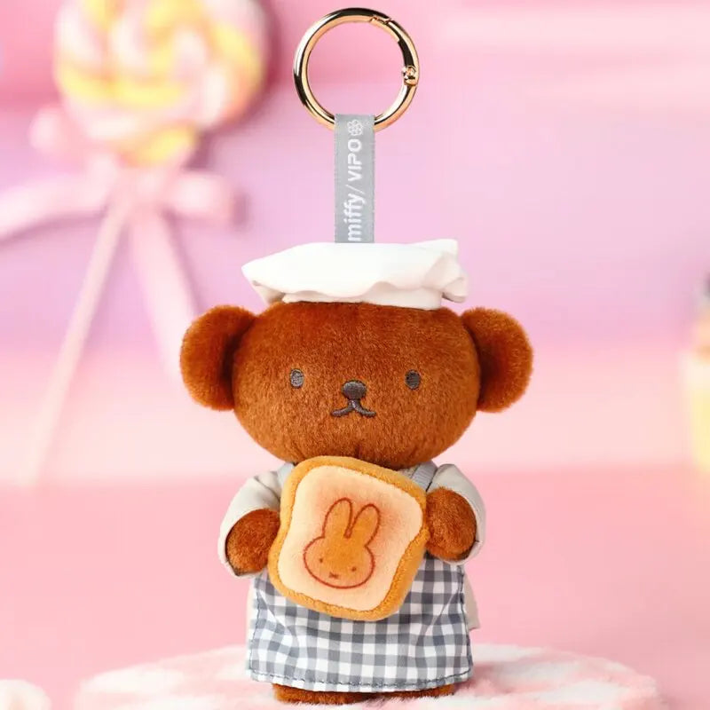 A brown teddy bear plush toy dressed as a baker with a checkered apron and a layered bread item, attached to a gold keyring.