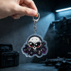 A hand holds an anime-inspired acrylic keychain charm featuring a skull and guns against a dark background.