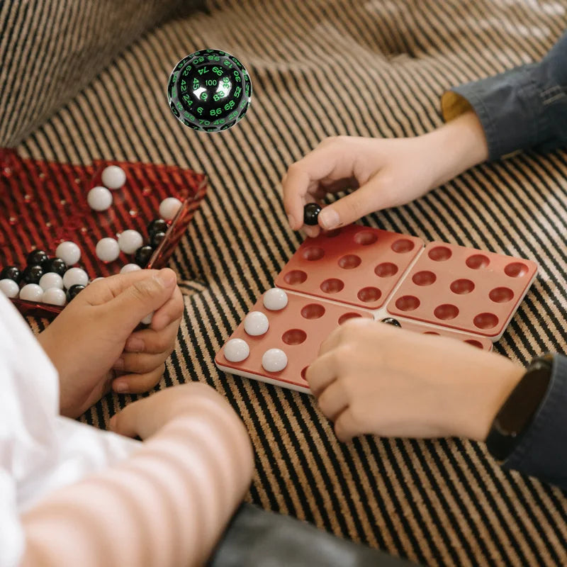 Hands are playing with a large 100-sided acrylic polyhedral die and small white and red beads on a striped fabric.