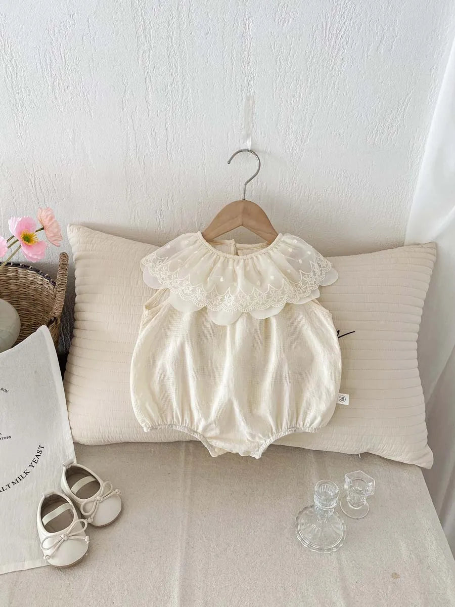 A cream-colored baby girl sleeveless jumpsuit with a lace collar hanging on a hanger against a white wall.