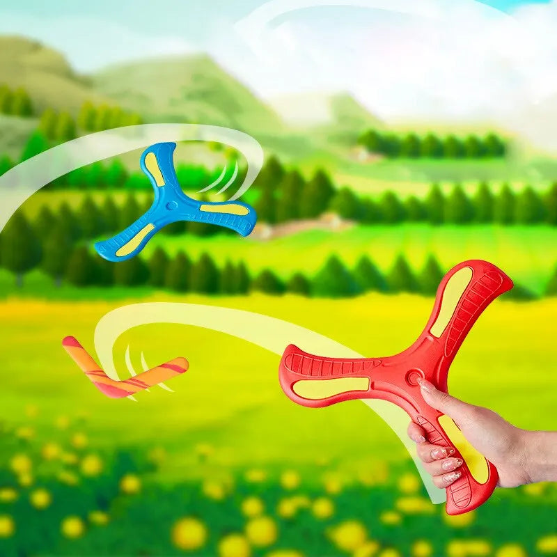 A hand holds a red soft foam boomerang flying disc against a bright green outdoor landscape.